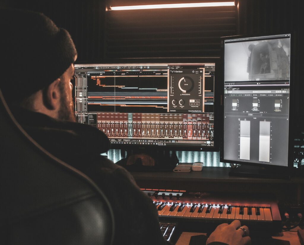 Music producer working in a studio, focused on multiple computer screens displaying sound editing software and audio tracks.