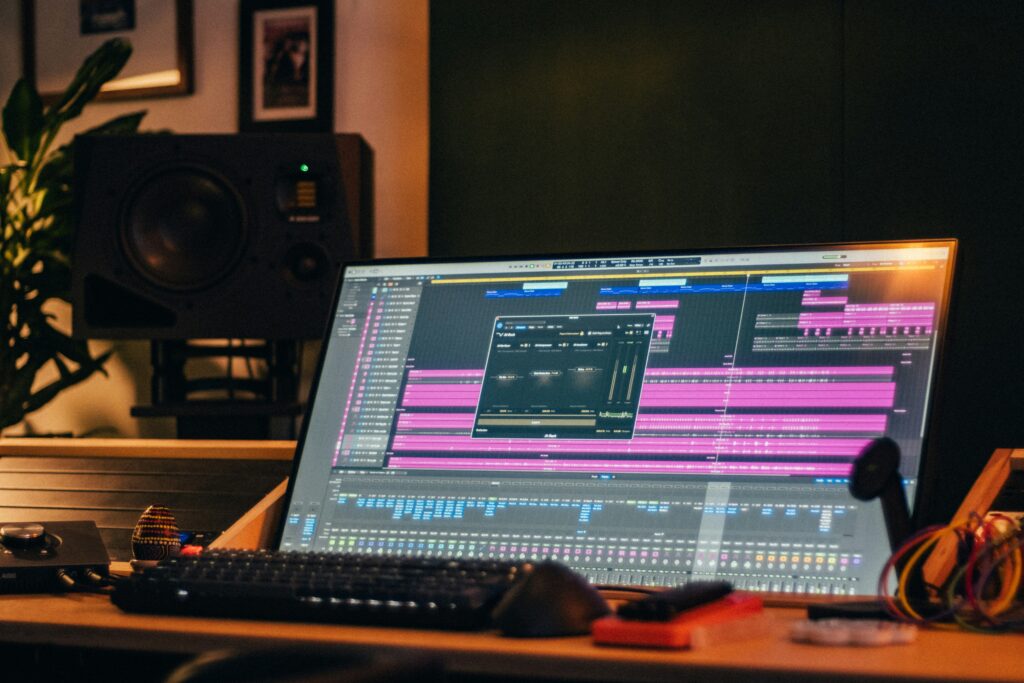 Music production workspace featuring a large monitor displaying a digital audio workstation with colorful audio tracks. A studio speaker and plants are visible in the background.